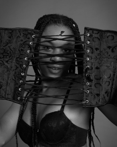 Caleann in black sixties-style bra and girdle, looking thorugh the lases of a black underbust corset
